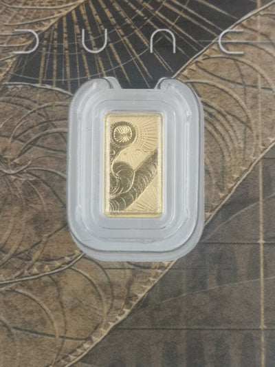 A gold bar with an engraved sand worm design, housed in a clear protective plastic capsule, set against a textured brown and beige background with the film name 'Dune'.