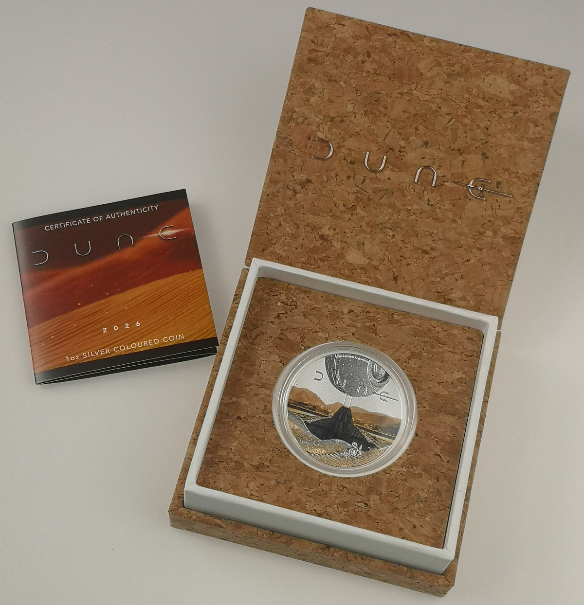 A silver-colored coin from the movie Dune, displayed inside an open cork-lined presentation box with a white insert. A certificate of authenticity booklet is placed to the left on a light gray background.