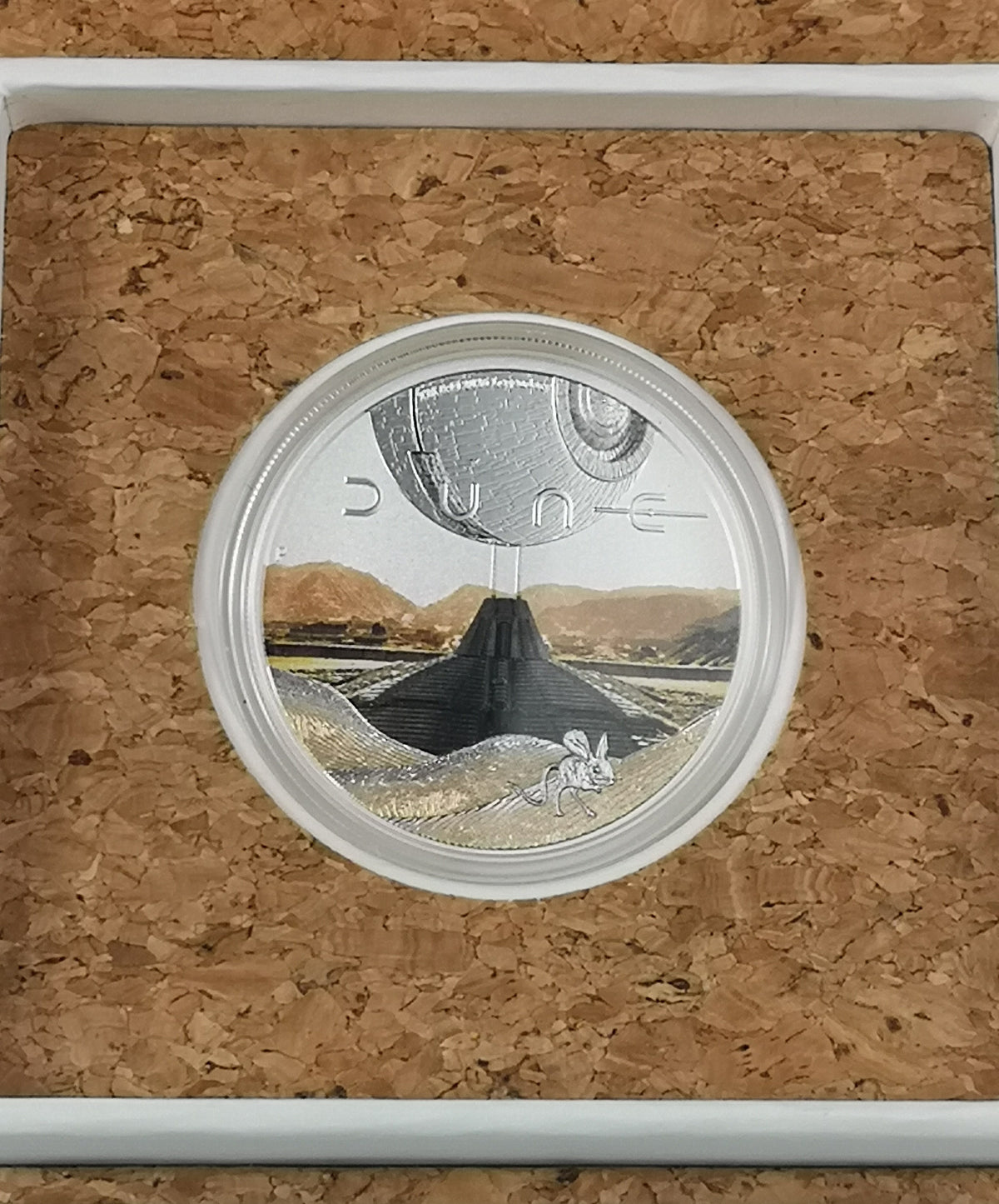 A Dune collectible coin featuring a desert landscape with a pyramid-like structure, a white mouse, and a metallic dome, displayed inside a white frame on a cork background. The word 'DUNE' is visible on the coin.